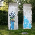 Berliner Mauer Zwei Segmente der Berliner Mauer mit Graffiti eines Cowboys auf einer grünen Wiese, umgeben von Bäumen.