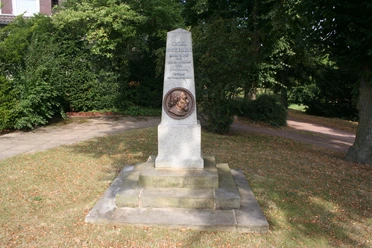 Ein Steinmonument mit Büste, umgeben von Bäumen in einem Park, ehrt eine historische Figur.