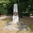 Ein Steinmonument mit Büste, umgeben von Bäumen in einem Park, ehrt eine historische Figur.
