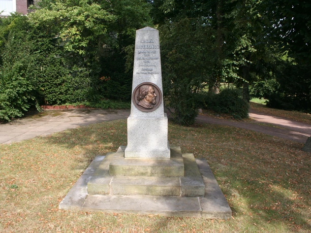 Ein Steinmonument mit Büste, umgeben von Bäumen in einem Park, ehrt eine historische Figur.