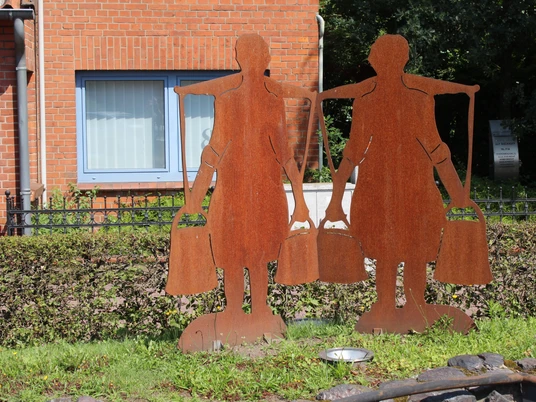 Dörverden Melkerinnen-Denkmal Metallskulptur von zwei Melkerinnen, die Eimer tragen, vor einem roten Backsteingebäude.