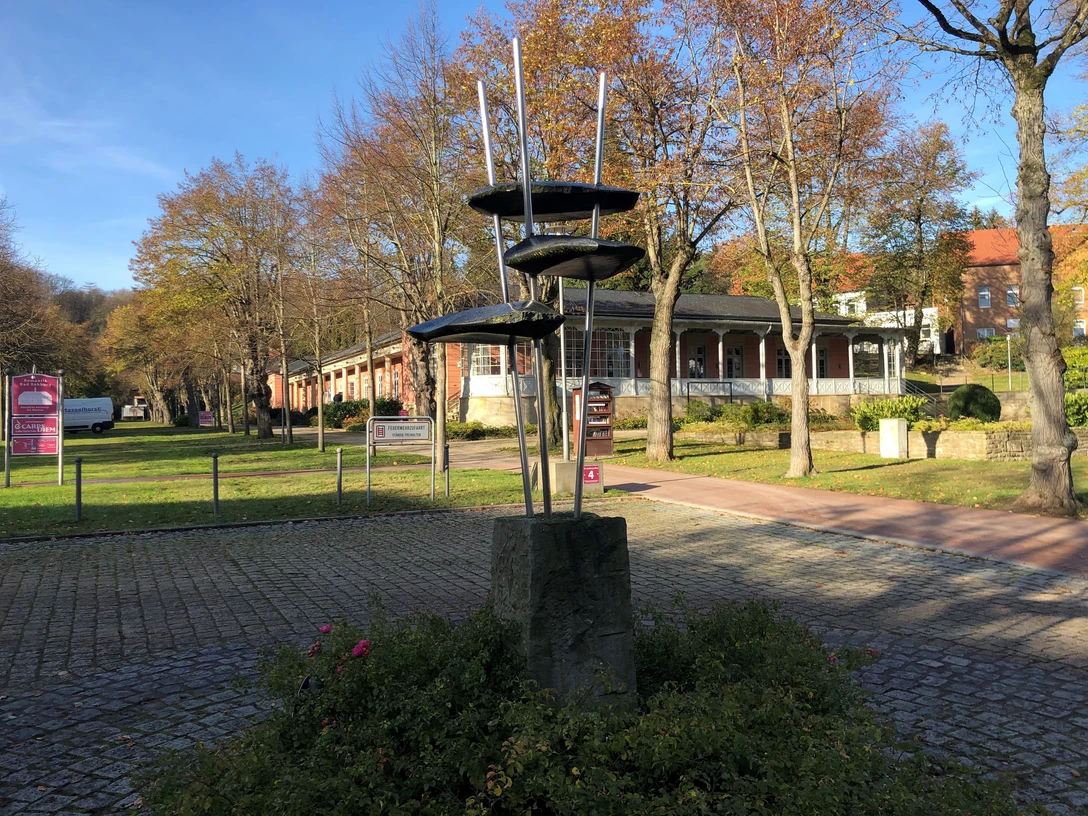 Skulptur "Juvenil" Stählerne Skulptur auf einem Sockel in herbstlicher Parkanlage, umgeben von Bäumen und Gebäuden.