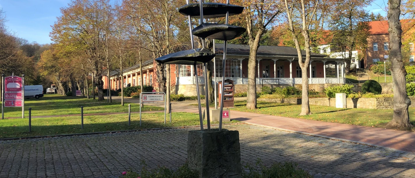 Skulptur "Juvenil" Stählerne Skulptur auf einem Sockel in herbstlicher Parkanlage, umgeben von Bäumen und Gebäuden.
