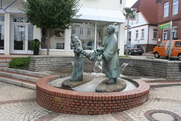 Brunnen-Skulptur einer Frau, die Wäsche auswringt, inmitten eines gepflasterten Platzes mit Bäumen.