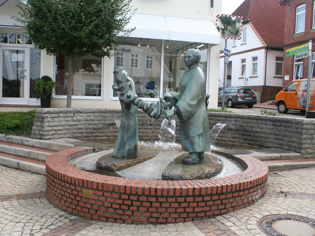 Brunnen-Skulptur einer Frau, die Wäsche auswringt, inmitten eines gepflasterten Platzes mit Bäumen.