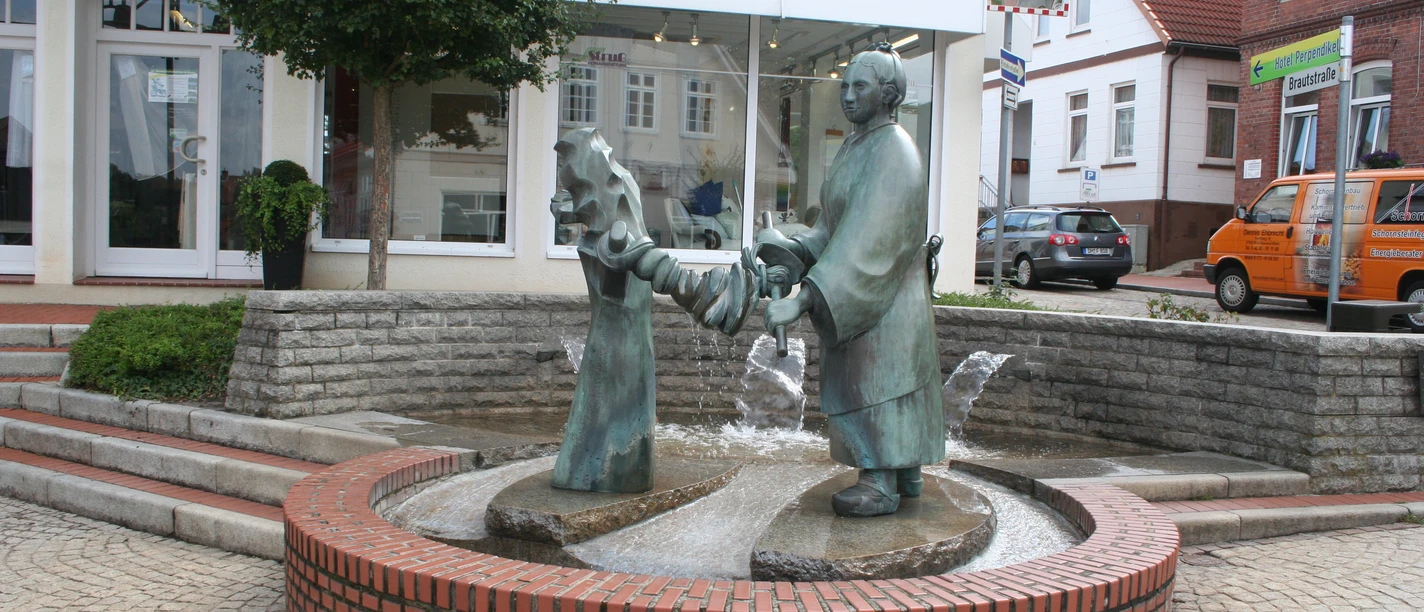 Brunnen-Skulptur einer Frau, die Wäsche auswringt, inmitten eines gepflasterten Platzes mit Bäumen.