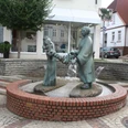 Brunnen - Die Wringerin Brunnen-Skulptur einer Frau, die Wäsche auswringt, inmitten eines gepflasterten Platzes mit Bäumen.