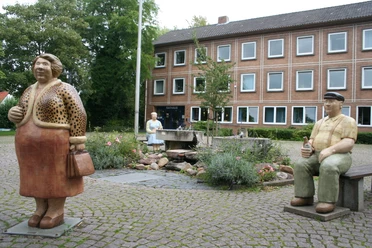 Rathaus Petershagen mit Lechner Figuren Bronzefiguren von Menschen vor dem Rathaus Petershagen, umgeben von einem kleinen Brunnen und bunten Blumen.