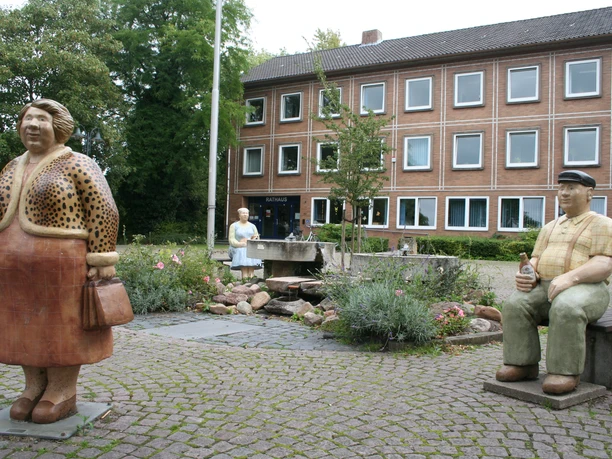 Rathaus Petershagen mit Lechner Figuren Bronzefiguren von Menschen vor dem Rathaus Petershagen, umgeben von einem kleinen Brunnen und bunten Blumen.