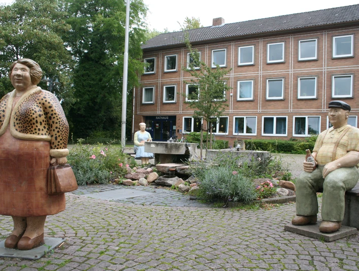 Rathaus Petershagen mit Lechner Figuren Bronzefiguren von Menschen vor dem Rathaus Petershagen, umgeben von einem kleinen Brunnen und bunten Blumen.