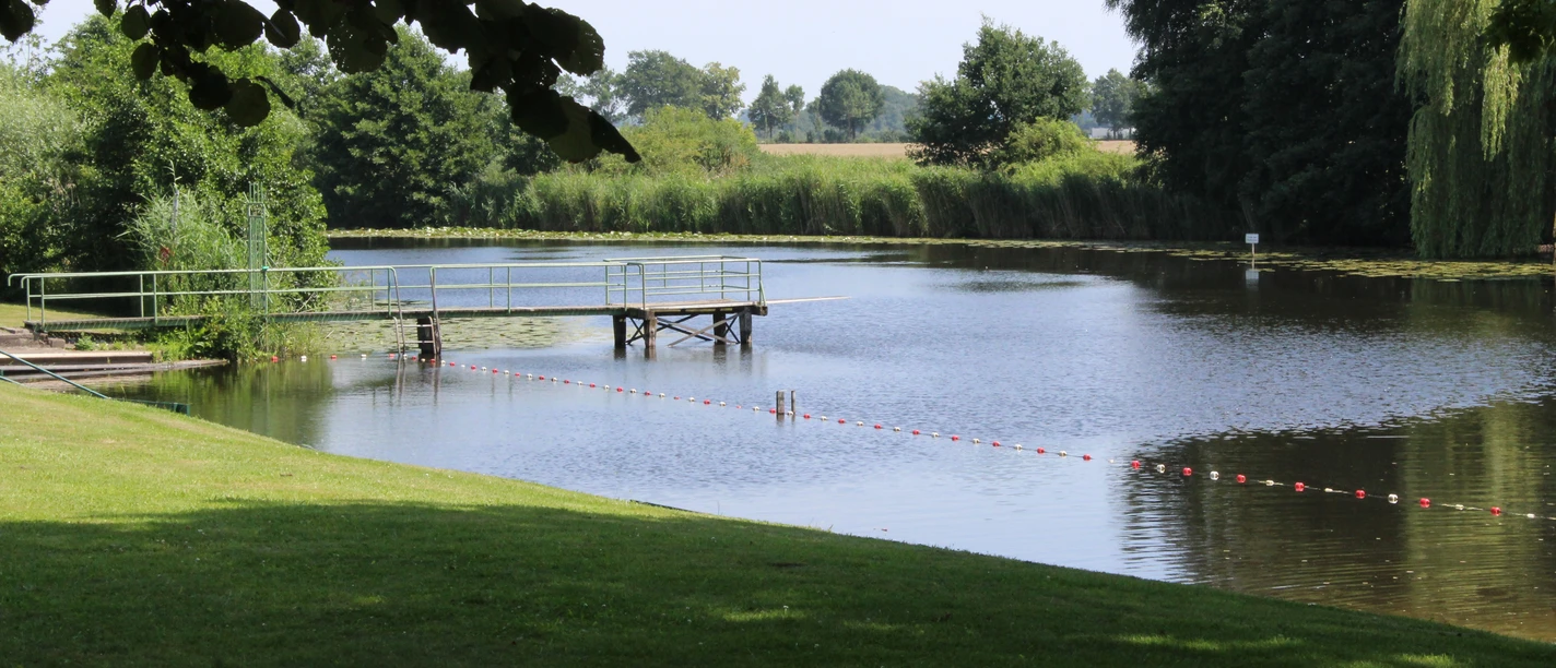 Badestelle am Blender See mit Steg, umgeben von grüner Wiese und Bäumen, lädt zum Entspannen ein.