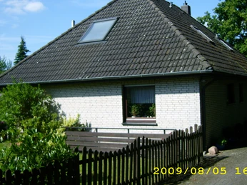 Eingebettet in eine grüne Umgebung steht ein weiß verklinkertes Haus mit schrägem Dach und Holzzaun.