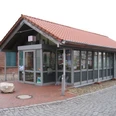 Kleines Tourismusbüro mit rotem Ziegeldach und Glasfenstern, umgeben von gepflasterten Flächen.Small tourist office with red tiled roof and glass windows, surrounded by paved areas.Lille turistkontor med rødt tegltag og glasvinduer, omgivet af asfalterede områder.Klein VVV-kantoor met rood pannendak en glazen ramen, omgeven door geplaveide ruimtes.