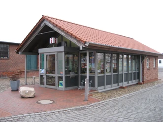 TourismusService Bruchhausen-Vilsen Kleines Tourismusbüro mit rotem Ziegeldach und Glasfenstern, umgeben von gepflasterten Flächen.