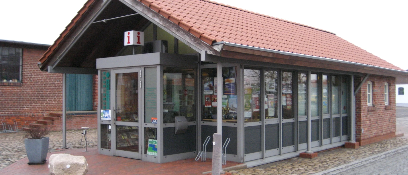 TourismusService Bruchhausen-Vilsen Kleines Tourismusbüro mit rotem Ziegeldach und Glasfenstern, umgeben von gepflasterten Flächen.