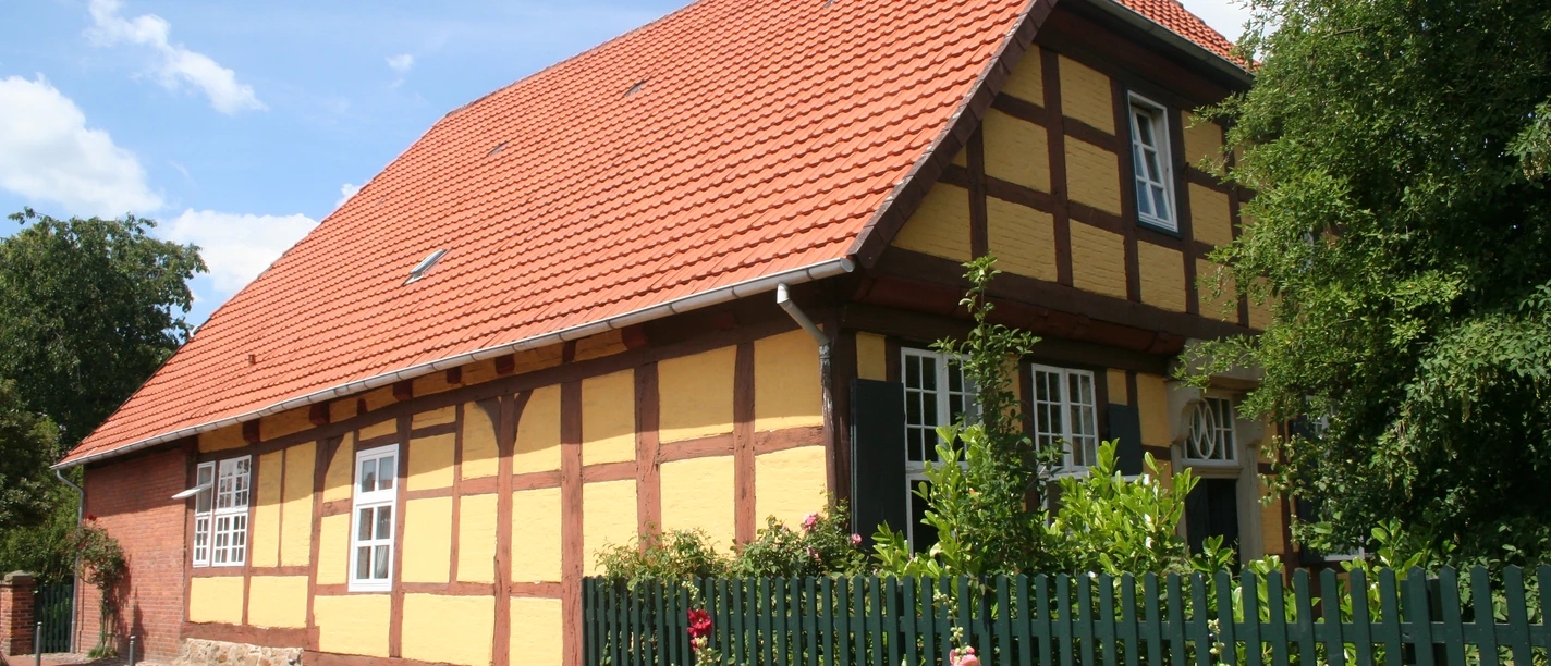 Fachwerkhaus mit roten Ziegeln, umgeben von Rosensträuchern und grünem Zaun unter blauem Himmel.