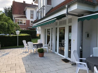 Terasse und Eingang Sonnige Terrasse mit weißen Stühlen und Tischen vor einem Gebäude mit großen Glasfenstern.