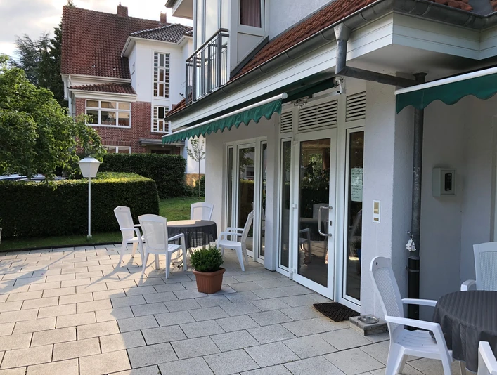 Terasse und Eingang Sonnige Terrasse mit weißen Stühlen und Tischen vor einem Gebäude mit großen Glasfenstern.