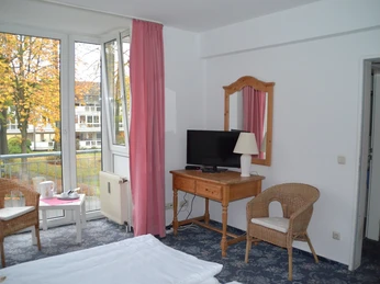 Blick aus dem Zimmer Ein Hotelzimmer mit zwei Korbsesseln, Tisch, Fernseher und Fenster mit Blick auf herbstliche Bäume.