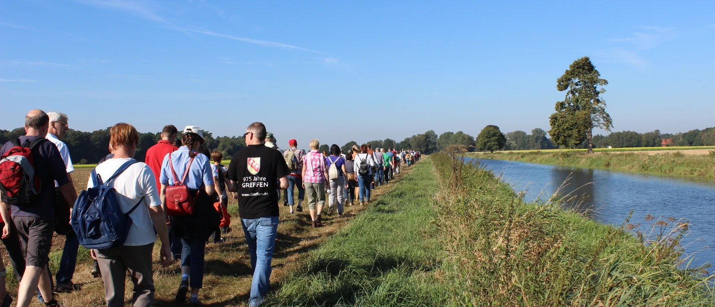 Die Ems in Greffen (Harsewinkel) Eine große Wandergruppe spaziert entlang eines Kanals an einem sonnigen Tag, flankiert von grüner Natur.