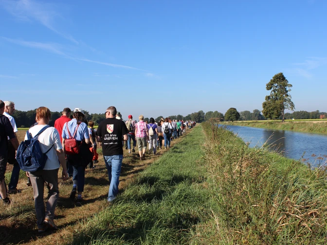 Die Ems in Greffen (Harsewinkel) Eine große Wandergruppe spaziert entlang eines Kanals an einem sonnigen Tag, flankiert von grüner Natur.