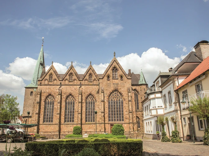 Außenansicht einer gotischen Kirche mit hohen Fenstern, umliegenden Gebäuden und grünem Garten.Exterior view of a Gothic church with high windows, surrounding buildings and a green garden.Udsigt til en gotisk kirke med høje vinduer, omkringliggende bygninger og en grøn have.Buitenaanzicht van een gotische kerk met hoge ramen, omringende gebouwen en een groene tuin.
