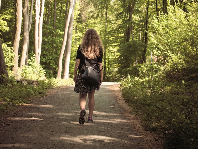 Eine Person mit langen Haaren geht auf einem Waldweg im Sonnenlicht spazieren.A person with long hair is walking along a forest path in the sunlight.En person med langt hår går langs en skovsti i sollyset.Een persoon met lang haar loopt over een bospad in het zonlicht.