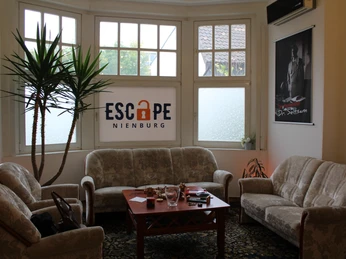 Escape Nienburg Ein einladender Aufenthaltsraum mit Polstermöbeln und einem zentralen Holztisch, Pflanzen im Hintergrund.An inviting lounge with upholstered furniture and a central wooden table, plants in the background.En indbydende lounge med polstrede møbler og et centralt træbord, planter i baggrunden.Een uitnodigende lounge met gestoffeerde meubels en een centrale houten tafel, planten op de achtergrond.