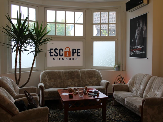 Escape Nienburg Ein einladender Aufenthaltsraum mit Polstermöbeln und einem zentralen Holztisch, Pflanzen im Hintergrund.An inviting lounge with upholstered furniture and a central wooden table, plants in the background.En indbydende lounge med polstrede møbler og et centralt træbord, planter i baggrunden.Een uitnodigende lounge met gestoffeerde meubels en een centrale houten tafel, planten op de achtergrond.