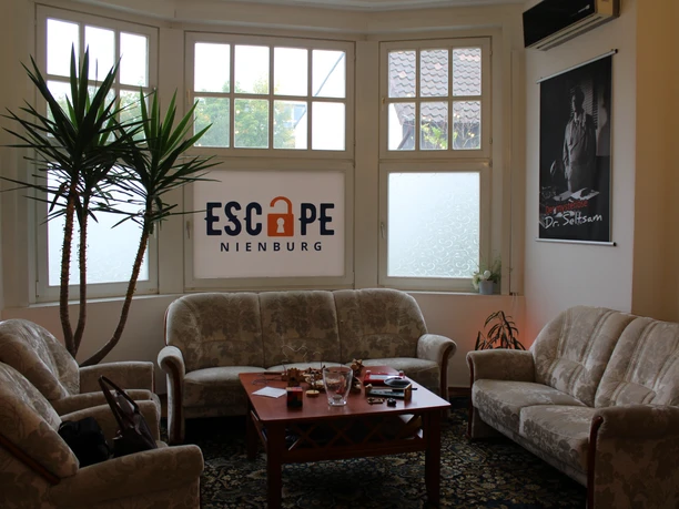 Escape Nienburg An inviting lounge with upholstered furniture and a central wooden table, plants in the background.