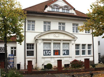 Historisches Gebäude in Nienburg, weiß gestrichen, mit Escape-Room-Logo in den Fenstern. Historic building in Nienburg, painted white, with Escape Room logo in the windows. Historisk bygning i Nienburg, hvidmalet, med escape room-logo i vinduerne. Historisch gebouw in Nienburg, wit geschilderd, met escape room-logo in de ramen.