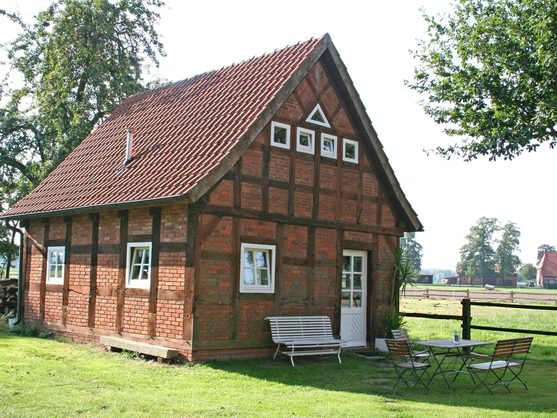 Fachwerkhaus auf einem grünen Hof mit Gartenmöbeln, umgeben von Bäumen und ländlicher Landschaft.