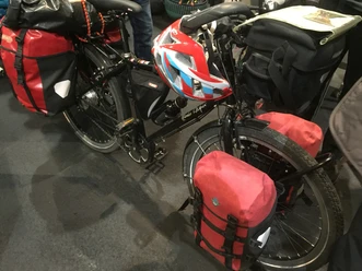 Ein schwarzes Touringrad mit roter Fahrradtasche und Helm steht gepackt auf einem Boden aus grauem Asphalt.