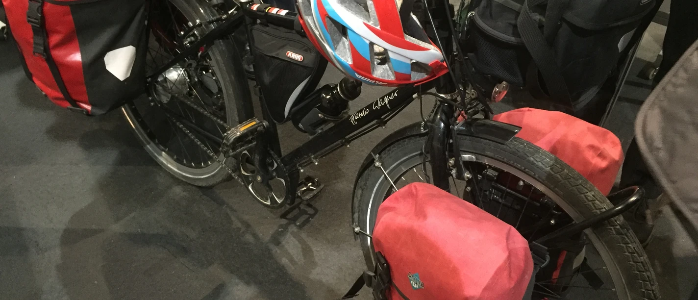 Ein schwarzes Touringrad mit roter Fahrradtasche und Helm steht gepackt auf einem Boden aus grauem Asphalt.