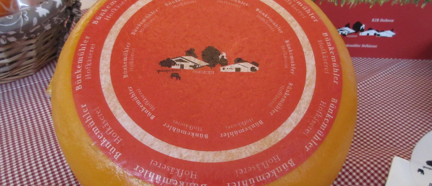 Bünkemühler Hofkäserei Großer, runder Laib Käse mit rotem Etikett der Bünkemühler Hofkäserei auf karierter Tischdecke.