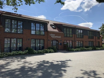 Backsteinfassade des Leister Aparthotels mit großen Fenstern und gepflastertem Vorplatz unter blauem Himmel.