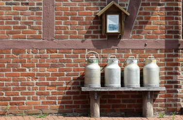 Milchkannen Vier traditionelle Milchkannen stehen auf einer Holzbank vor einer Backsteinmauer mit Fachwerk.