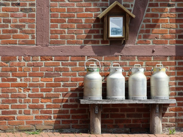 Milchkannen Vier traditionelle Milchkannen stehen auf einer Holzbank vor einer Backsteinmauer mit Fachwerk.