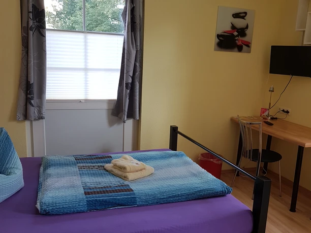 A bright room with a bed, blue and purple bed linen, a window alcove and a desk.