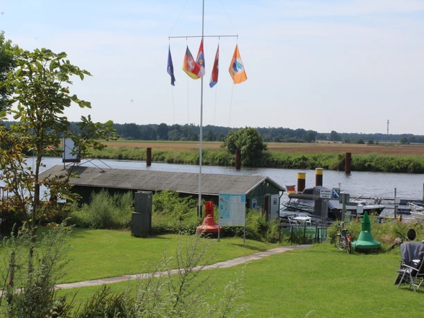 Grüne Flusslandschaft mit Bootssteg, Fahnenmasten und Hütten im Wassersportverein Palm Beach Dörverden.