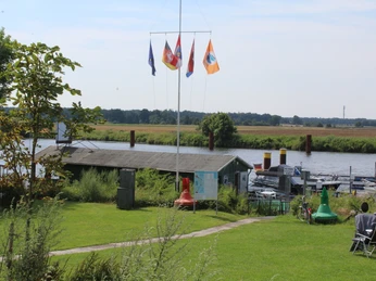 Wassersportverein Palm Beach Dörverden Grüne Flusslandschaft mit Bootssteg, Fahnenmasten und Hütten im Wassersportverein Palm Beach Dörverden.