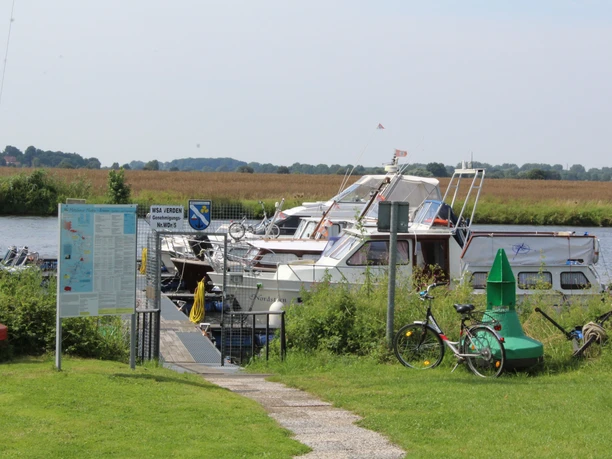 Boote liegen am Steg vor einer sanften Flusslandschaft; ein Fahrrad lehnt am Wegesrand.