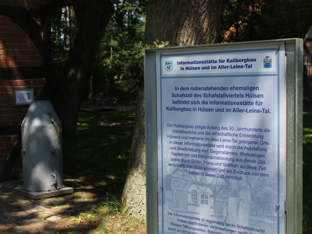 Dörverden Kalibergbau Hülsen Informationstafel zum Kalibergbau in Hülsen, umgeben von Bäumen und einem Ziegelgebäude im Hintergrund.