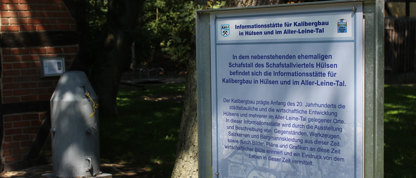 Dörverden Kalibergbau Hülsen Informationstafel zum Kalibergbau in Hülsen, umgeben von Bäumen und einem Ziegelgebäude im Hintergrund.