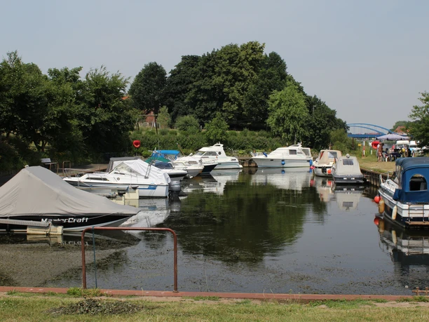 Eine ruhige Wasserfläche mit vertäuten Sportbooten im Vordergrund, umgeben von grünen Bäumen.