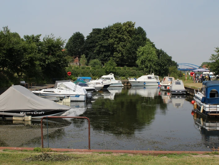 Eine ruhige Wasserfläche mit vertäuten Sportbooten im Vordergrund, umgeben von grünen Bäumen.