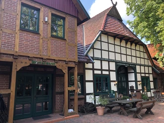 Half-timbered house with red roof tiles, green-framed windows and restaurant lettering, surrounded by trees.