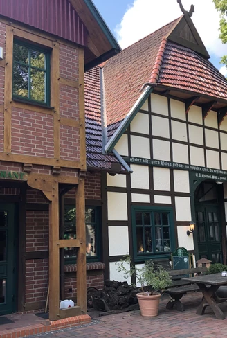 Fachwerkhaus mit roten Dachziegeln, grünumrahmten Fenstern und Restaurant-Schriftzug, umgeben von Bäumen.Half-timbered house with red roof tiles, green-framed windows and restaurant lettering, surrounded by trees.Bindingsværkshus med røde tagsten, vinduer med grønne rammer og restaurantbogstaver, omgivet af træer.Vakwerkhuis met rode dakpannen, ramen met groene kozijnen en restaurantbelettering, omringd door bomen.