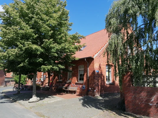 Rotes Backsteingebäude mit Tradition, umgeben von Bäumen, blauer Himmel im Hintergrund, Fahrräder seitlich.