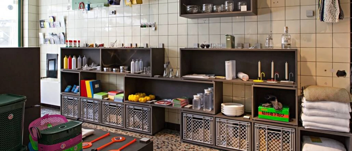 Utensil Shelves are attached to the tiled wall in a high-quality interior design store. Various colorful household utensils, towels and decorative baskets are arranged on the shelves and on a wooden cabinet.